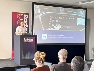 Gwendolin Niehues speaks on stage at Radiodays Europe next to a screen showing a car dashboard with a digital display