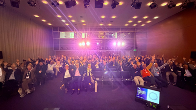 The audience for the WorldDAB Conference Session at IBC2026 - a conference room full of people with their hands in the air waving at the photographer on the stage