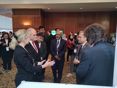 WorldDAB Project Director Bernie O'Neill stands on the left talking to Malaysian Minister of Communications and Digital, Fahmi Fadzil in front of the WorldDAB pavilion stand. Other delegates and photographers stand around them in a large hotel conference hall.