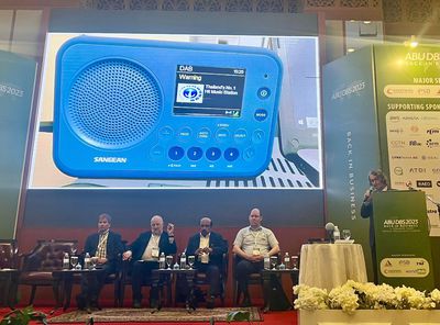 Dr Les Sabel is on a stage speaking at a lectern, in front of a screen showing a photo of a blue DAB digital radio. The four other panellists in the conference sit on the stage below the screen.