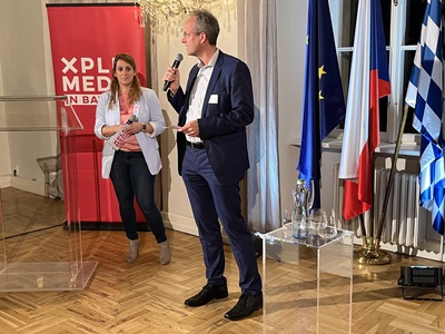 Melanie Walter stands to the left in front of a red banner with the logo of XPLR: Media in Baveria. She is looking at Dr. Thorsten Schmiege speaking holding a microphone and standing in front of EU, Czech and Bavarian flags in a window. 