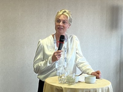 Jacqueline Bierhorst holds a microphone standing behind a round high drinks table while speaking to the WorldDAB happy hour reception