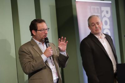 Joe D'Angelo stands on stage to the left, speaking into a microphone he's holding, while Desmond S. Fuller stands to the right.