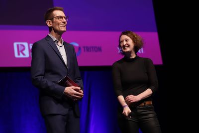 Paul Robinson stands on stage on the left speaking, with Edita Kudlácova on the right looking at him smiling